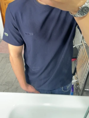 Mörkblå t-shirt från Lacoste - Säljer en mörkblå t-shirt från Lacoste med klassisk rund hals och diskret logotyp på bröstet. T-shirten har korta ärmar och är gjord i mjuk bomull. Den ikoniska krokodilen finns broderad på ena ärmen. Perfekt för en avslappnad och stilren look.