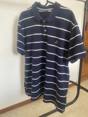 Randig blå polotopp från Gant - Säljer en klassisk marinblå polotopp från Gant med vita horisontella ränder och krage. Toppen har korta ärmar och knappar framtill. Perfekt för dig som gillar en sportig och stilren look.