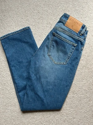 Blå raka jeans från Filippa K, Stella  - Säljer ett par helt nya klassiska blå jeans från Filippa K med rak passform och hög midja. Jeansen har fickor, bälteshällor och en snygg läderpatch med logga bak i midjan. Tillverkade i slitstark denim som ger en tidlös look. Modell Stella, cropped. 