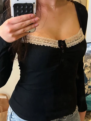 Svart långärmad topp med spets - Svart långärmad topp med dekorativ beige spetskant vid urringningen och knappar framtill. Toppen har en figurnära passform och är tillverkad i ett ribbat material som ger en snygg struktur. Perfekt för dig som gillar feminina detaljer.