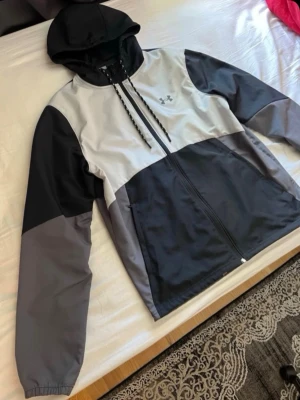 Under Armour vindjacka med huva, möts bara upp i Stockholm  - Snygg vindjacka från Under Armour i svart, grått,vitt och ljusgrått. Jackan har hel dragkedja, två sidofickor och justerbar huva med snören. Perfekt för blåsiga dagar och sportiga outfits. Materialet är lätt och vindtätt.