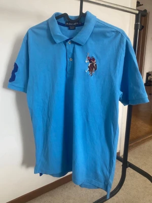 Blå piké från  US POLO ASSN - Säljer en klassisk blå pikétröja från US POLO ASSN med den ikoniska polospelaren broderad på bröstet och en stor siffra på ärmen. Tröjan har korta ärmar, krage och knappar framtill. Perfekt för en avslappnad men ändå stilren look.