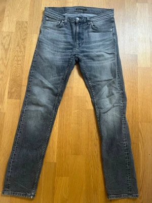 Grå slim fit jeans från Nudie Jeans - Säljer ett par grå slim fit jeans från Nudie Jeans med snygga slitningar och klassiska femficksdetaljer. Jeansen har en cool tvättad look och är tillverkade i bomull. Perfekt för dig som gillar en avslappnad och trendig stil. Storleken är 28W/30L och modellen Lean Dean