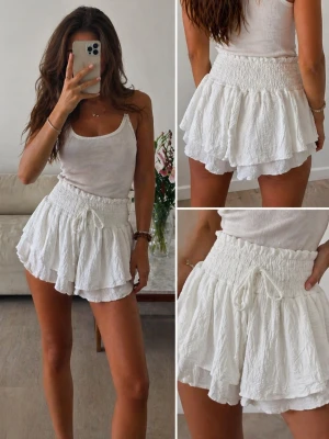 Vita volangshorts med snörning - Säljer ett par vita shorts med volang och hög smockad midja. De har snörning framtill och är tillverkade i ett lätt, krusat bomullstyg som ger en luftig känsla. Perfekta för varma dagar och en avslappnad stil. Aldrig använda, de har bara legat i lådan. 