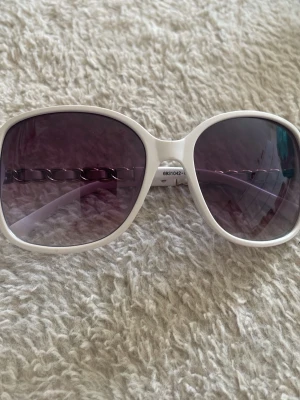 Vita solglasögon med kedjedetalj - Stora, vita solglasögon med silverfärgad kedjedetalj på bågarna. Glasen är mörka med en lätt toning som ger en cool look. Perfekt accessoar för att lyfta din stil under soliga dagar. Bågarna är i syntetmaterial och har en trendig, oversized form.