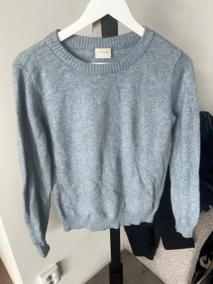 Ljusblå stickad tröja från VILA - Ljusblå stickad tröja från VILA med rund halsringning och ribbade muddar vid ärmslut och nederkant. Enkel och klassisk design som är perfekt för lager-på-lager. Materialet är mjukt och behagligt, vilket gör den riktigt skön att bära.