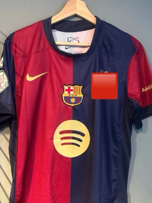 Barcelona matchtröja Nike Lamine Yamal - Säljer en Barcelona matchtröja från Nike med Lamine Yamal och nummer 19 på ryggen. Tröjan är röd och blå med klubbmärke, sponsorlogga och detaljer i gult. Materialet är lätt och andas, perfekt för fotboll eller träning.