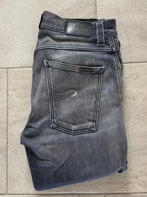 Grå jeans från Nudie Jeans - Säljer ett par grå jeans från Nudie Jeans med klassisk femficksdesign och snygga kontrastsömmar på bakfickorna. Jeansen har en rak passform och är tillverkade i denim med cool tvättad look. Perfekt för dig som gillar stilrena och trendiga jeans.