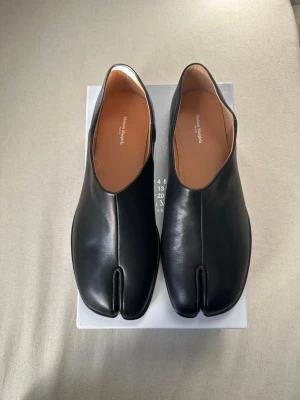 Maison Margiela Tabis - Unika svarta Tabi loafers från Maison Margiela i slätt läder. Skorna har den ikoniska delade tån och en minimalistisk design med låg klack. Insidan är fodrad i brunt läder för extra komfort. Perfekt för dig som gillar statement-plagg och modern stil.