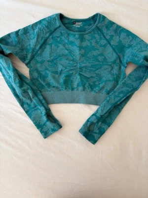 Turkos cropad träningströja M - Säljer en turkos cropad träningströja med mönstrad design. Tröjan har långa ärmar med tumhål och är tillverkad i 90% polyamid och 10% elastan för extra stretch och komfort. Perfekt för gymmet eller yoga.