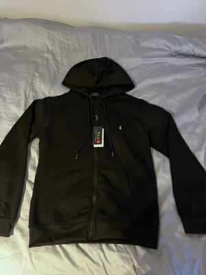 Svart hoodie från Polo Ralph Lauren - Snygg svart hoodie från Polo Ralph Lauren med dragkedja framtill och klassisk huva med dragsko. Ikoniska vita loggan broderad på bröstet. Tillverkad i mjukt bomullsmaterial och har känguruficka. Perfekt för en chill och stilren look.