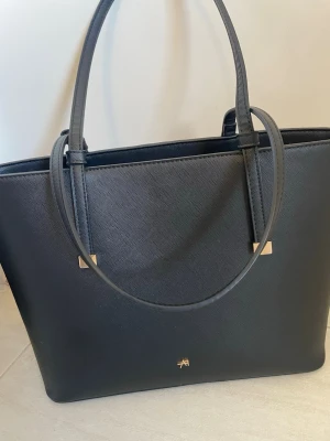 Svart handväska från Anna Field - Stilren svart handväska med två långa axelband och guldfärgade detaljer. Väskan har en klassisk rektangulär form och är tillverkad i ett strukturerat syntetmaterial. Diskret logotyp framtill och enkel design som passar till många outfits.