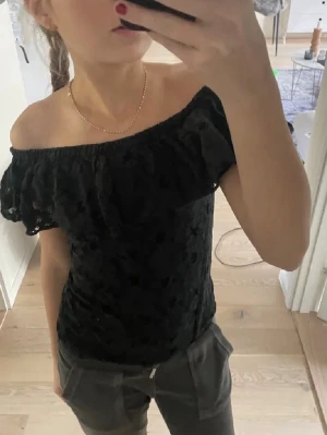 Svart offshoulder blus med spets  - Säljer en svart offshoulder blus med volang och spetsdetaljer. Blusen har en romantisk känsla och är perfekt för dig som gillar att sticka ut. Den är ärmlös och har en tight passform som framhäver axlarna.