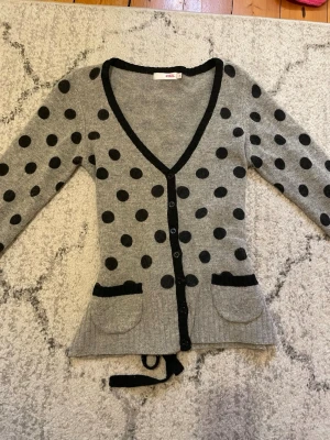 Grå kofta med svarta prickar från ZOUL - Snygg grå kofta med svarta prickar från ZOUL. Koftan har djup V-ringning, svarta kanter, två fickor framtill och knytband i midjan. Långa ärmar och knäppning med svarta knappar. Perfekt för dig som gillar lekfulla mönster och detaljer. Pris går alltid att diskutera!! Mer som en S då den är justerbar