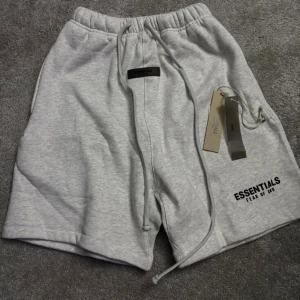 Grå Essentials shorts med dragsko - Säljer ett par grå shorts från Essentials Fear of God med elastisk midja och dragsko. De har en avslappnad passform och logotypen tryckt på ena benet. Perfekta för en chill look och tillverkade i mjukt bomullsmaterial. Helt nya, fraktar inom 24 timmar