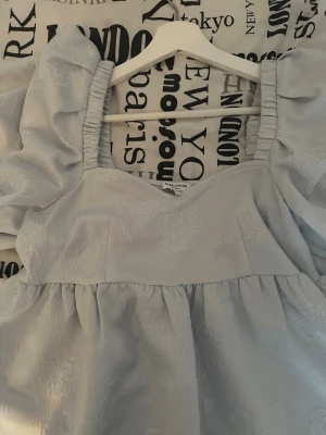 Ljusblå klänning med puffärmar - Ljusblå klänning från Bubbleroom med puffiga kortärmar och subtilt blommigt mönster i tyget. Klänningen har en v-ringad front.