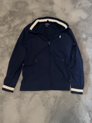 Marinblå vindjacka från Polo Ralph Lauren - Snygg marinblå vindjacka från Polo Ralph Lauren med vit logga på bröstet. Jackan har dragkedja framtill, två fickor med dragkedja och vita detaljer vid krage och ärmslut. Perfekt för en sportig och stilren look.