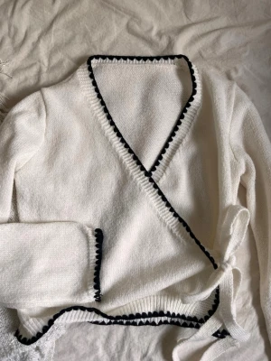 Vit omlottkofta med svart kant - Säljer en vit stickad omlottkofta med svarta dekorativa kanter och knytband i sidan. Koftan har lång ärm och ribbstickad nederkant. Perfekt för dig som gillar en clean och stilren look med en twist.