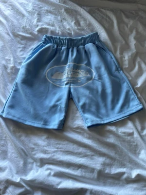 Ljusblå shorts med tryck - Säljer ett par ljusblå shorts med resår i midjan och ett vitt tryck framtill som föreställer ett landskap. Shortsen har en avslappnad passform och är perfekta för varma dagar. Materialet känns mjukt och bekvämt.