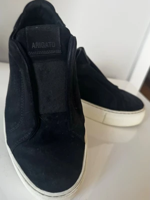 Svarta mocka sneakers dice laced från Axel Arigato - Säljer ett par svarta sneakers från Axel Arigato i stl 39 med clean design och bred vit sula. Skorna är gjorda i mocka och har dice laced, elastiskt band över tungan för en snygg och enkel look. Perfekta för dig som gillar minimalistisk stil.