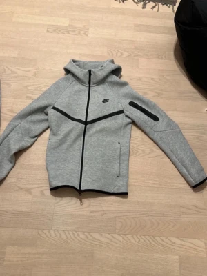 Nike tech hoodie - Säljer en grå hoodie från Nike med svarta detaljer och dragkedja framtill. Hoodien har en huva, svarta linjer över bröstet och armen samt en ficka med dragkedja på ärmen. Perfekt för dig som gillar sportig stil.