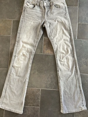 Grå raka jeans med fickdetaljer - Snygga grå jeans med raka ben och klassisk femficksdesign. Baksidan har två fickor med lock och knapp. Jeansen har normal midja och är tillverkade i denim med en lätt tvättad look. Perfekta för en avslappnad och trendig stil.