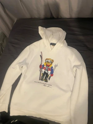 Vit hoodie Polo Bear från Ralph Lauren - Vit hoodie från Ralph Lauren med ikoniskt Polo Bear-motiv på framsidan. Björnen är klädd i skidkläder och håller skidor, vilket ger ett sportigt och lekfullt intryck. Hoodien har huva och är tillverkad i mjukt material som känns skönt mot huden.
