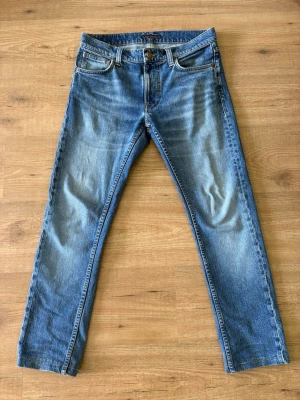 Blå jeans från Nudie Jeans - Säljer ett par klassiska blå jeans från Nudie Jeans med raka ben och femficksdesign. Jeansen har snygga slitningar och kontrastsömmar, samt läderpatch på bakfickan. Materialet är denim i bomull och passformen är avslappnad. De är också uppsydda men det är bara att släpa på sömen så blir de 2 cm längre/fabriks längd. 