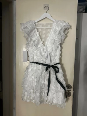 Vit volangklänning från Bubbleroom - Supersöt vit kortklänning från Bubbleroom med volanger, v-ringning och blommigt strukturmönster. Klänningen har ett svart knytband i midjan (man kan ta bort om man vill) och är ärmlös. Perfekt till studenten! Ny med prislapp