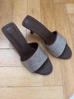 Helt nya från NA-KD - Säljer ett par bruna pumps från NA-KD med öppen tå och slip-in modell. Skorna har en bred asymmetrisk rem täckt av glittrande strass och en smal klack på ca 5 cm. Endast testade. 