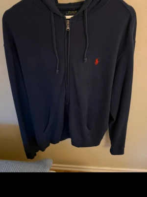 Mörkblå hoodie från Polo Ralph Lauren - Mörkblå hoodie med dragkedja från Polo Ralph Lauren. Klassisk design med huva och snören, fickor framtill och den ikoniska röda logon broderad på bröstet. Perfekt för en avslappnad och stilren look.