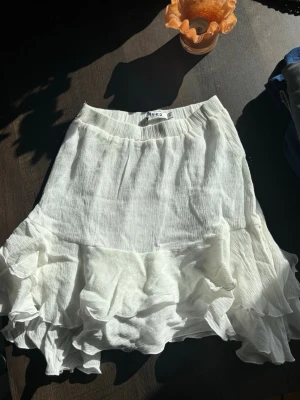 Vit volangkjol från NA-KD 34 - Supersöt vit kortkjol från NA-KD med volangdetaljer och lätt skrynkligt tyg. Kjolen har en flowy och luftig känsla, perfekt för varma dagar. Elastisk midja och flera lager volanger som ger en cool och trendig look.