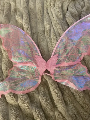Rosa fjärilsvingar med glitter - Säljer ett par stora fjärilsvingar med band i rosa med glittriga detaljer och skimrande material. Vingarna har ett genomskinligt tyg med rosa glittermönster och en liten fjäril i mitten. Perfekt för maskerad eller cosplay som fe eller älva.