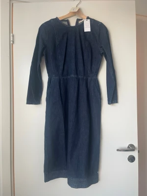 jeansklänning med lång ärm - Snygg långärmad jeansklänning i mörkblå denim med markerad midja och rund halsringning. Klänningen har en rak, lång siluett och dragkedja i ryggen. Perfekt för dig som gillar en stilren och tidlös look.