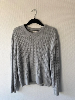 Gant tröja  - Säljer en snygg grå kabelstickad tröja från Gant med rund hals och diskret logga broderad på bröstet. Tröjan har långa ärmar och ribbade muddar vid ärmslut och nederkant. Perfekt för lager-på-lager och chill stil.