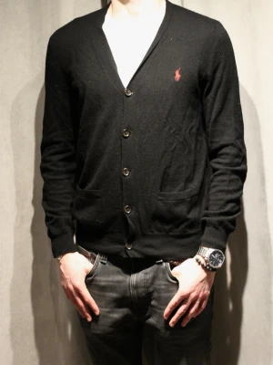 Svart kofta från Polo Ralph Lauren - Svart kofta med knappar från Polo Ralph Lauren. Klassisk design med två fickor framtill och röd broderad logga på bröstet. V-ringad och långärmad, perfekt att slänga på över en t-shirt. Materialet är mjukt och tunt, vilket gör den enkel att bära.