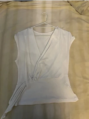 Vit topp - Snygg vit omlottopp med knyt i sidan och v-ringad hals. Ärmlös design som ger en clean och modern look. Perfekt att matcha med jeans eller kjol för en stilren outfit.