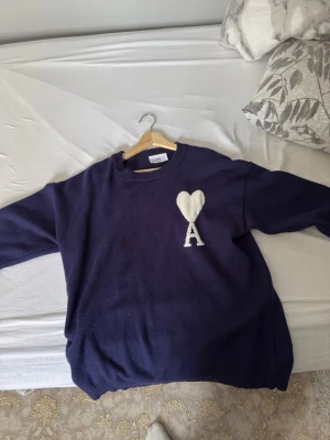 Mörkblå stickad tröja från Ami - Säljer en mörkblå stickad tröja från Ami med ett vitt hjärta och bokstaven 'A' broderat på bröstet. Tröjan har rund halsringning och ribbstickade muddar vid ärmslut och nederkant. Perfekt för att hålla sig varm och snygg.