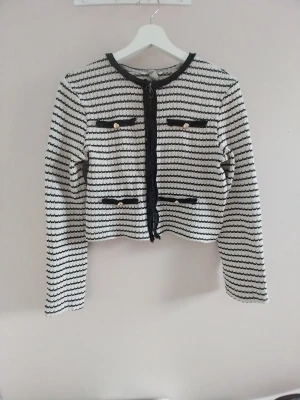 Randig svartvit kofta med gulddetaljer - Snygg svart/beige kofta med horisontella ränder och dekorativa guldfärgade knappar på fyra fakefickor. Koftan har rund halsringning, långa ärmar och svarta kanter längs öppningen och fickorna. Perfekt för en chic och trendig look. Sälger pga att jag inte använder och den är lite för kort på mig