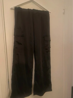 Svarta cargobyxor med vida ben - Säljer ett par svarta satin cargobyxor med bred passform och stora sidofickor. Byxorna har en avslappnad look och är tillverkade i ett glansigt material som ger en cool känsla. Perfekta för dig som gillar streetstyle och vill ha något bekvämt och trendigt.