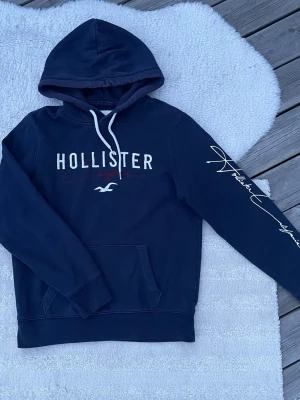 Mörkblå hoodie från Hollister - Mörkblå hoodie från Hollister med vit logotyp och röd signatur på bröstet. Hoodien har vit snörning i huvan, långärmad design och en stor ficka framtill. Materialet är mjuk bomull och polyester, perfekt för chill dagar. Snyggt tryck på ena ärmen. Lite solblekt vid huvan