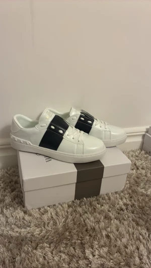 Vita Valentino sneakers med svart band - Snygga vita sneakers från Valentino med ett brett svart band över vristen och silverfärgade nitar. Skorna har en klassisk låg profil och är tillverkade i slätt läder med vit gummisula. Perfekta för dig som gillar stilrena och exklusiva detaljer.
