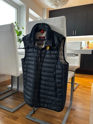 Mörkblå dunväst från Parajumpers - Snygg svart dunväst från Parajumpers med hög krage och den iconiska gula detaljen vid dragkedjan. Västen har två sidofickor med dragkedja för att hålla sakerna kvar. Den är använd en del men inga tecken på användning. Inga slitningar, hål eller liknande. Västen håller dig varm utan att bli för tung. Vid frågor är det bara att kontakta mig!