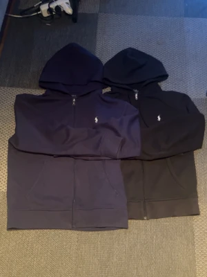 Svart och marinblå hoodie Polo Ralph Lauren - Två stilrena hoodies från Polo Ralph Lauren, en i svart och en i marinblått. Båda har dragkedja framtill, huva och den klassiska broderade loggan på bröstet. Tillverkade i mjukt bomullsmaterial och har fickor framtill. Perfekta för en avslappnad och snygg look. ‼️Ljuset är dåligt, inte tröjorna som tappar färg‼️
