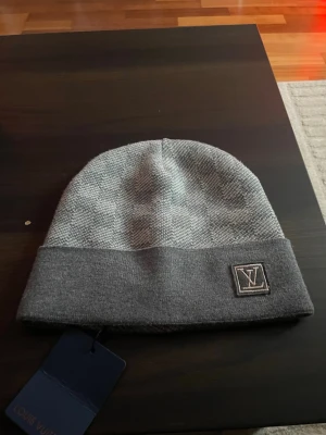 Grå mössa från Louis Vuitton - Snygg grå mössa från Louis Vuitton med uppvikt kant och diskret LV-logga framtill. Tillverkad i mjukt stickat material som känns lyxigt och bekvämt. Perfekt accessoar för att höja din stil under kyliga dagar.