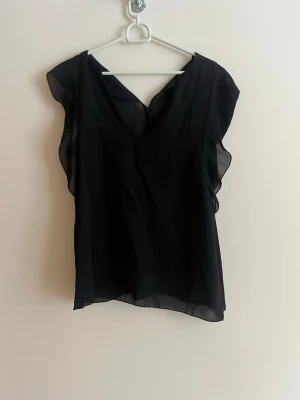 Svart blus med volangdetaljer - Snygg svart blus i luftig polyester med v-ringning och volangdetaljer längs ärmhålen. Blusen är ärmlös och har en lätt transparent känsla som ger en elegant look. Perfekt att styla med jeans eller kjol för en trendig outfit.