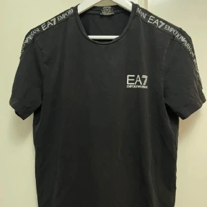 Svart EA7 Emporio Armani t-shirt - Snygg svart t-shirt från EA7 Emporio Armani med logga på bröstet och textdetaljer längs axlarna. Klassisk rund halsringning och korta ärmar. Perfekt för dig som gillar stilrena och trendiga plagg med en sportig touch.