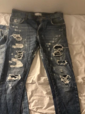 Blå slitna jeans med hål - Säljer ett par blå jeans med kraftiga slitningar och stora hål på både fram- och baksida. Jeansen har en klassisk femficksmodell och är tillverkade i denim. Perfekta för en edgy och avslappnad stil.