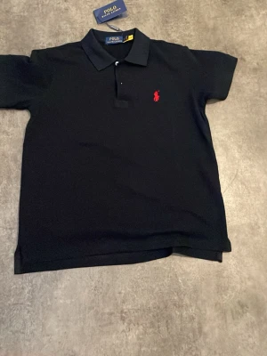 Svart pikétröja från Polo Ralph Lauren - Klassisk svart pikétröja från Polo Ralph Lauren med röd broderad logga på bröstet. Tröjan har krage, knappar framtill och korta ärmar. Perfekt för en stilren och avslappnad look.