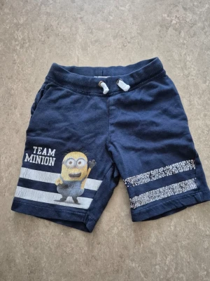 Shorts - Säljer ett par blå shorts med Minion-motiv och texten 'TEAM MINION' på ena benet. Shorts med snörning i midjan och vita ränder med mönster på benen. Perfekta för sommarens chill och häng.
